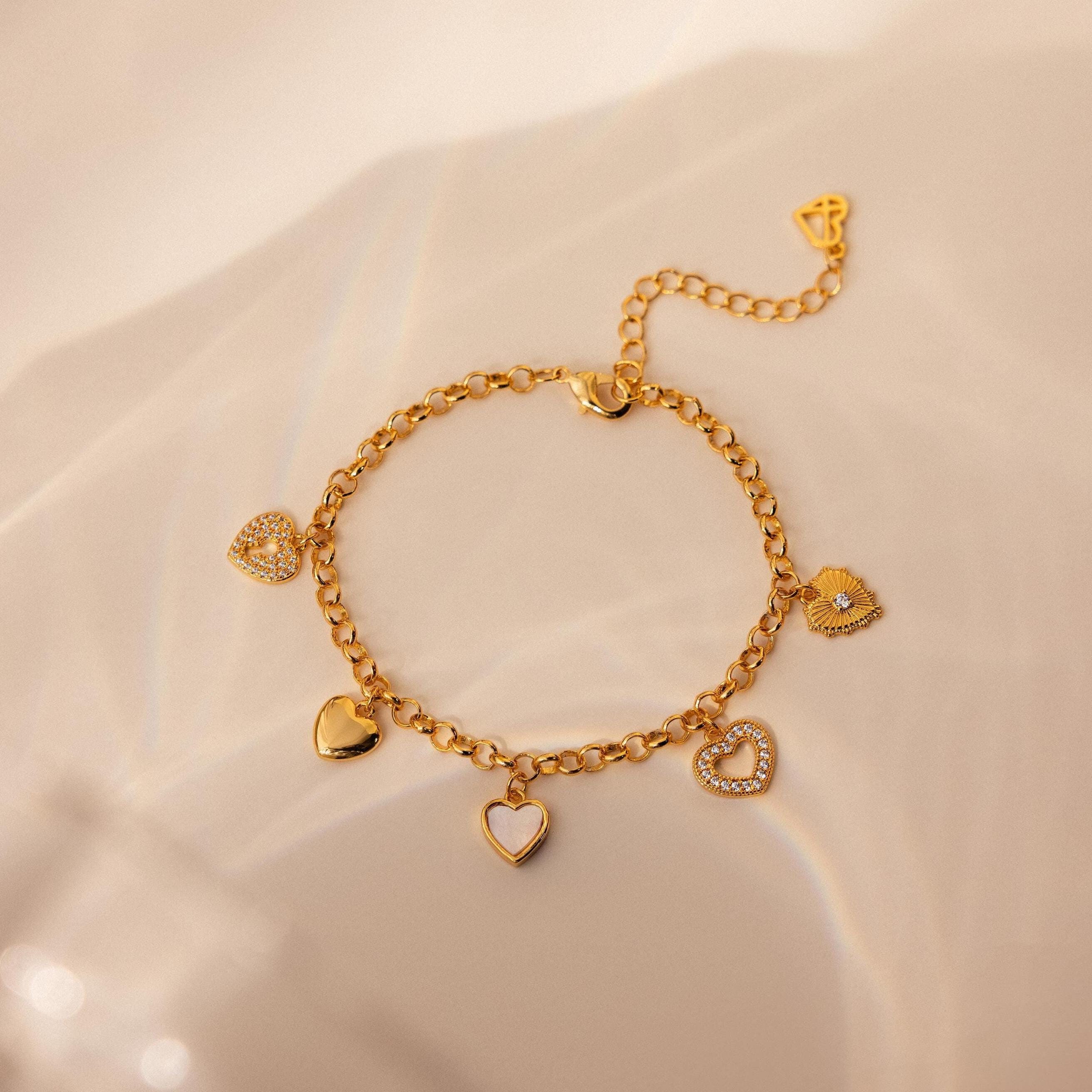 A gold chain charm bracelet with heart-shaped charms encrusted with sparkling CZ stones, displayed on a reflective surface.